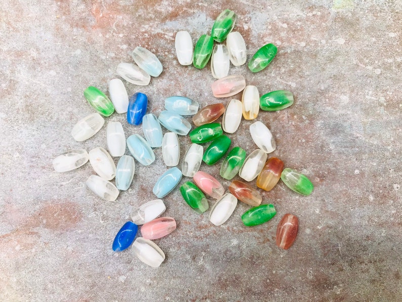 Pu&ograve; includere: Una collezione di perline di vetro ovali in vari colori, tra cui verde, blu, bianco e rosa. Le perline sono sparse su una superficie testurizzata.