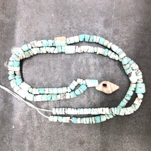 K&ouml;nnte beinhalten: Eine t&uuml;rkisfarbene und wei&szlig;e Perlenkette mit einem Holzanh&auml;nger. Die Kette besteht aus kleinen, unregelm&auml;&szlig;igen Perlen und ist auf einer wei&szlig;en Schnur aufgef&auml;delt.