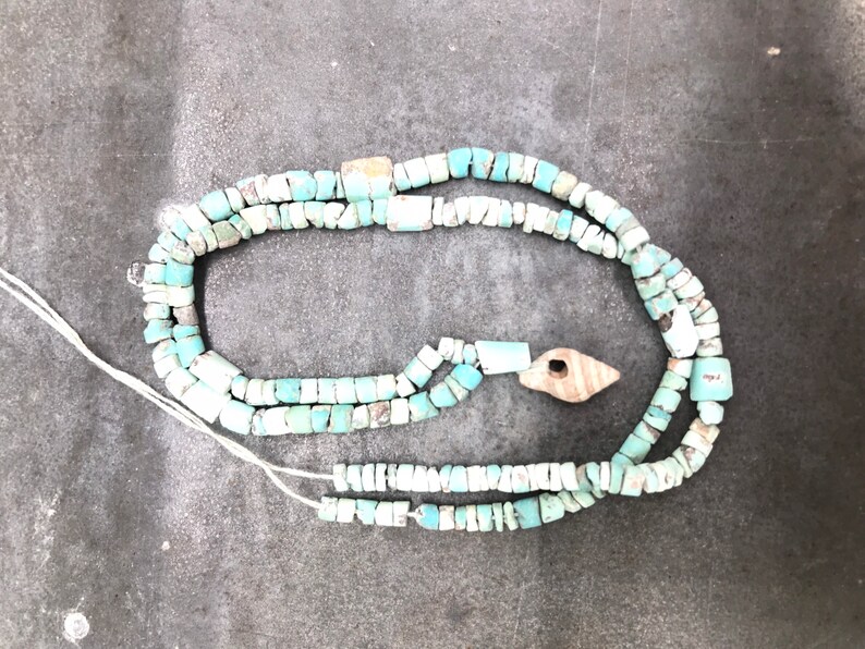 K&ouml;nnte beinhalten: Eine t&uuml;rkisfarbene und wei&szlig;e Perlenkette mit einem Holzanh&auml;nger. Die Kette besteht aus kleinen, unregelm&auml;&szlig;igen Perlen und ist auf einer wei&szlig;en Schnur aufgef&auml;delt.