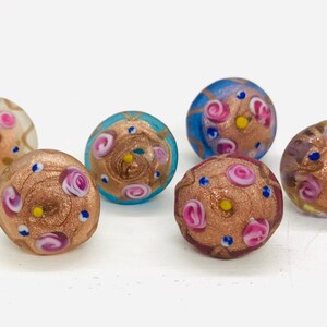 May include: Six small glass beads with a gold and pink swirl pattern. The beads are decorated with small pink and blue flowers and gold glitter.