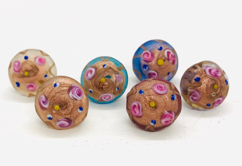 May include: Six small glass beads with a gold and pink swirl pattern. The beads are decorated with small pink and blue flowers and gold glitter.