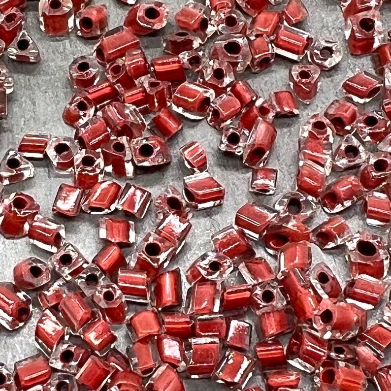 May include: A large quantity of small, red, square-shaped glass beads. The beads have a clear, transparent finish with a red hue.