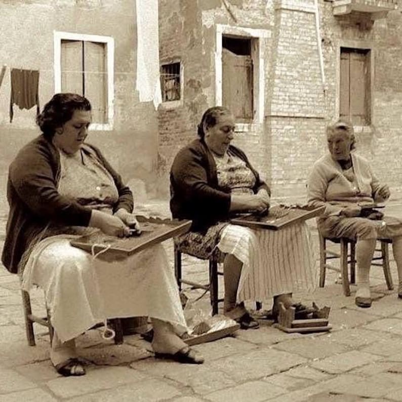 May include: Three women sit on chairs outside, working on a craft project. They are using their hands to manipulate small objects on a table in front of them. The women are wearing simple clothing and are focused on their work.