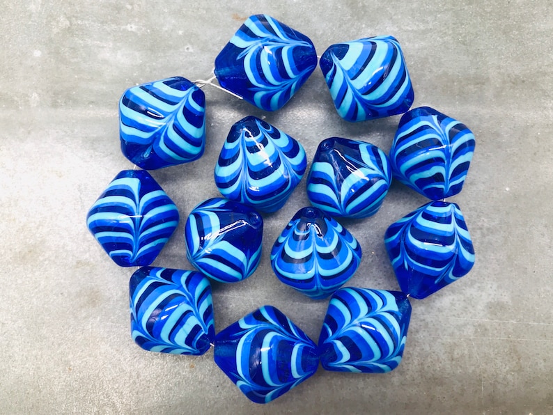 May include: A collection of 12 blue and white glass beads with a marbled swirl pattern. The beads are shaped like a diamond with rounded edges.