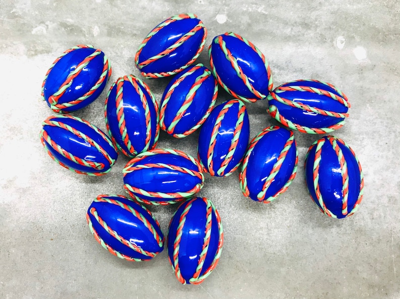 Op de afbeelding: Een groep van 12 blauwe ovale kralen met witte, groene en oranje strepen. De kralen liggen op een grijs oppervlak.