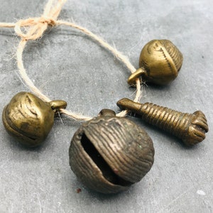 K&ouml;nnte beinhalten: Ein Satz aus vier antiken Messingglocken und einem kleinen Messing-Anh&auml;nger, die mit einer Schnur zusammengebunden sind. Die Glocken haben verschiedene Formen und Gr&ouml;&szlig;en und eine strukturierte Oberfl&auml;che.