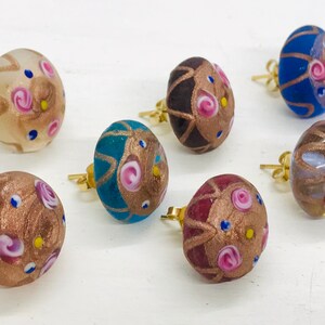 May include: Six pairs of colorful glass earrings with gold accents and floral designs. The earrings are round and have a textured surface.