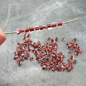 May include: A close-up of a pile of small, clear glass beads with a reddish-brown tint. The beads are scattered on a gray surface. A strand of the beads is strung on a gold wire.