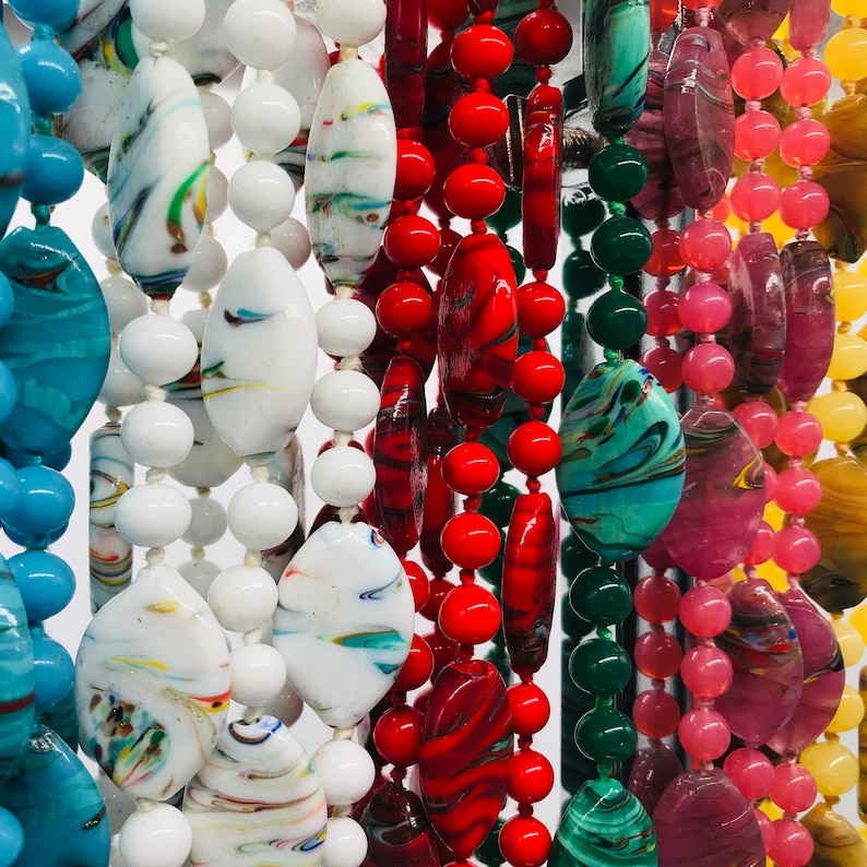 K&ouml;nnte beinhalten: Nahaufnahme von bunten Glasperlen, die zu Halsketten aufgereiht sind. Die Perlen haben verschiedene Formen und Gr&ouml;&szlig;en, darunter rund, oval und tropfenf&ouml;rmig. Die Farben umfassen Rot, Blau, Gr&uuml;n, Wei&szlig; und Pink.