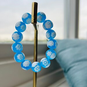 May include: A blue bracelet with white flower designs on each bead. The bracelet is displayed on a gold stand.