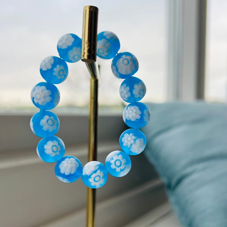 May include: A blue bracelet with white flower designs on each bead. The bracelet is displayed on a gold stand.