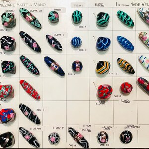May include: A display of handmade Venetian glass beads in various colors and patterns. The beads are arranged in rows on a white card with labels for each color and style. The card also includes the text "PERLE VENEZIANE FATTE A MANO" and "MADE VENETIAN BEADS".