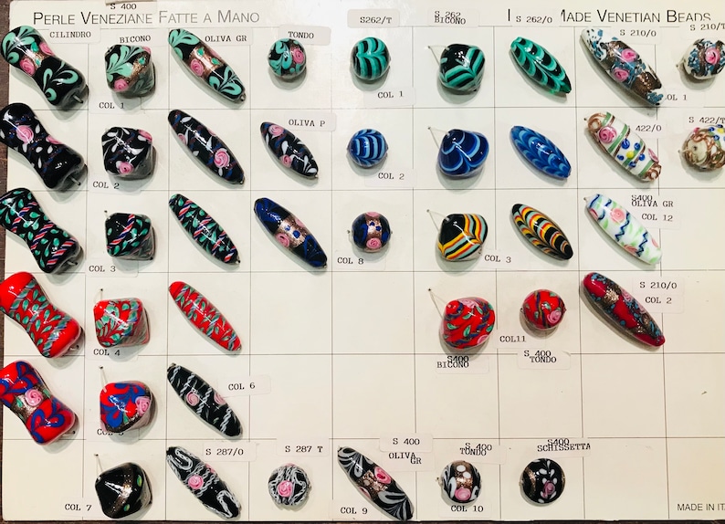 May include: A display of handmade Venetian glass beads in various colors and patterns. The beads are arranged in rows on a white card with labels for each color and style. The card also includes the text "PERLE VENEZIANE FATTE A MANO" and "MADE VENETIAN BEADS".