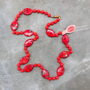 K&ouml;nnte beinhalten: Eine rot-schwarze Musterkette mit einem goldenen Verschluss und einem wei&szlig;en Etikett mit der Aufschrift "RMV".