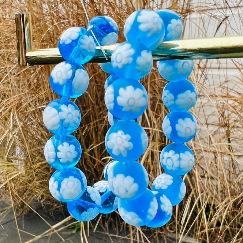 May include: Two blue glass bracelets with white flower designs. The bracelets are strung on a gold metal bar.