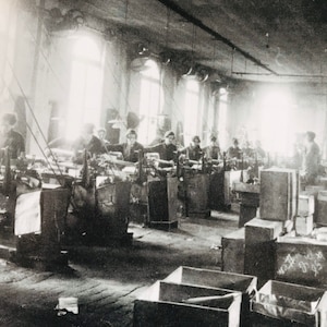 May include: Black and white photo of a factory interior with workers operating machinery. The workers are wearing clothing typical of the early 20th century. The factory is filled with large wooden crates.