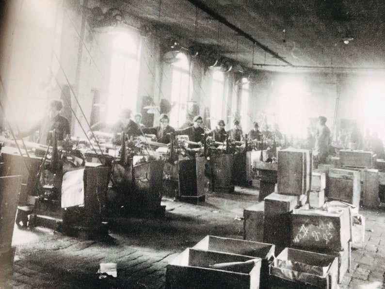 May include: Black and white photo of a factory interior with workers operating machinery. The workers are wearing clothing typical of the early 20th century. The factory is filled with large wooden crates.
