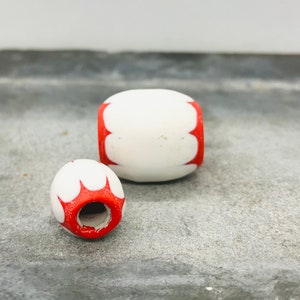 Può includere: Due perline di legno bianche con disegni dipinti in rosso. Le perline sono di forma ovale e hanno un foro al centro.