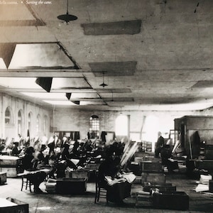 May include: Black and white photo of a large room with many people working at desks. The room has a high ceiling with skylights and is filled with wooden crates and stacks of paper. The text "Cerniuz della canna. Sorting the cane." is visible in the top left corner of the image.