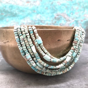 K&ouml;nnte beinhalten: Eine mehrreihige Halskette aus t&uuml;rkisfarbenen Perlen. Die Perlen sind unregelm&auml;&szlig;ig geformt und haben eine leicht raue Textur. Die Halskette wird in einer Schale aus Messing pr&auml;sentiert.