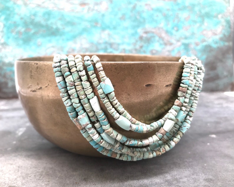 K&ouml;nnte beinhalten: Eine mehrreihige Halskette aus t&uuml;rkisfarbenen Perlen. Die Perlen sind unregelm&auml;&szlig;ig geformt und haben eine leicht raue Textur. Die Halskette wird in einer Schale aus Messing pr&auml;sentiert.