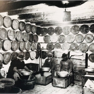 May include: Black and white photo of a room with four people sorting through a large quantity of round, woven baskets. The baskets are hanging on the walls and stacked on shelves. The people are wearing hats and are sitting at tables with bowls in front of them.