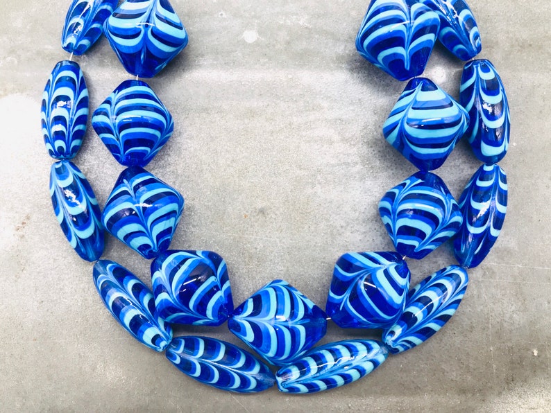 May include: A necklace made of blue and white glass beads. The beads are in a variety of shapes, including oval and square. The beads are all hand-painted with a swirling pattern.