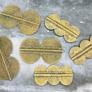 Può includere: Una collezione di abbellimenti in metallo color ottone. I pezzi presentano una serie di cerchi concentrici collegati da una barra centrale strutturata. I disegni variano per dimensioni e forma, alcuni hanno due cerchi e altri tre.