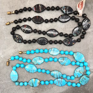 K&ouml;nnte beinhalten: Zwei Str&auml;nge bunter Glasperlenketten. Eine Kette ist schwarz mit wei&szlig;en Wirbeln und die andere ist t&uuml;rkis mit gelben und wei&szlig;en Wirbeln.