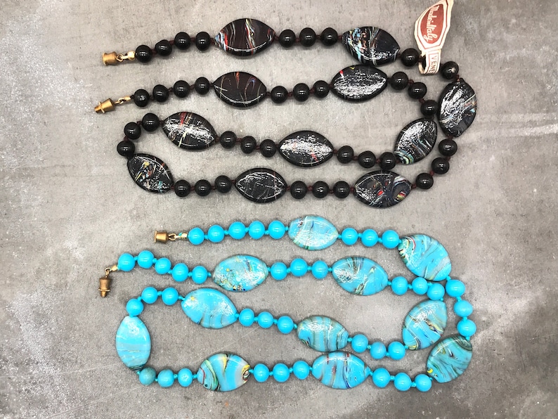 K&ouml;nnte beinhalten: Zwei Str&auml;nge bunter Glasperlenketten. Eine Kette ist schwarz mit wei&szlig;en Wirbeln und die andere ist t&uuml;rkis mit gelben und wei&szlig;en Wirbeln.