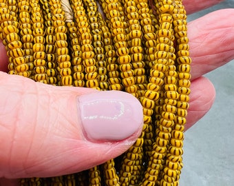May include: A close-up of a strand of yellow and brown striped glass beads. The beads are small and round, and they are strung together in a long strand.
