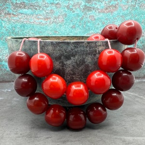 以下が含まれることがあります： ボウルに入っている赤と茶色の丸いビーズのネックレス。ビーズは滑らかで磨かれた素材で作られています。