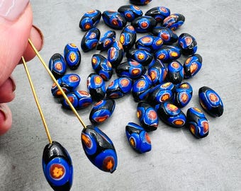 Puede incluir: Una colección de cuentas de vidrio negras y azules con acentos naranja y blanco. Las cuentas son de forma ovalada y tienen un patrón moteado.
