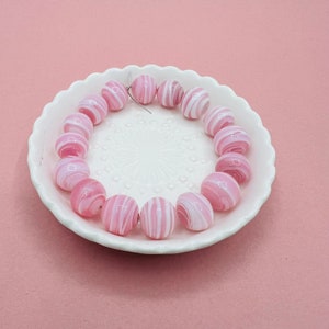 May include: A strand of pink and white striped glass beads on a white plate. The beads are round and have a glossy finish.