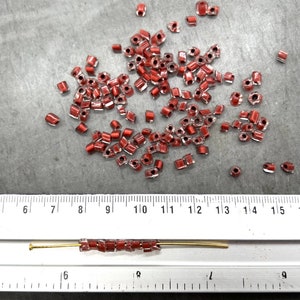 May include: A close-up of a ruler measuring 15 inches, with a line of small, red, square-shaped glass beads strung on a gold wire. The beads are scattered around the ruler.