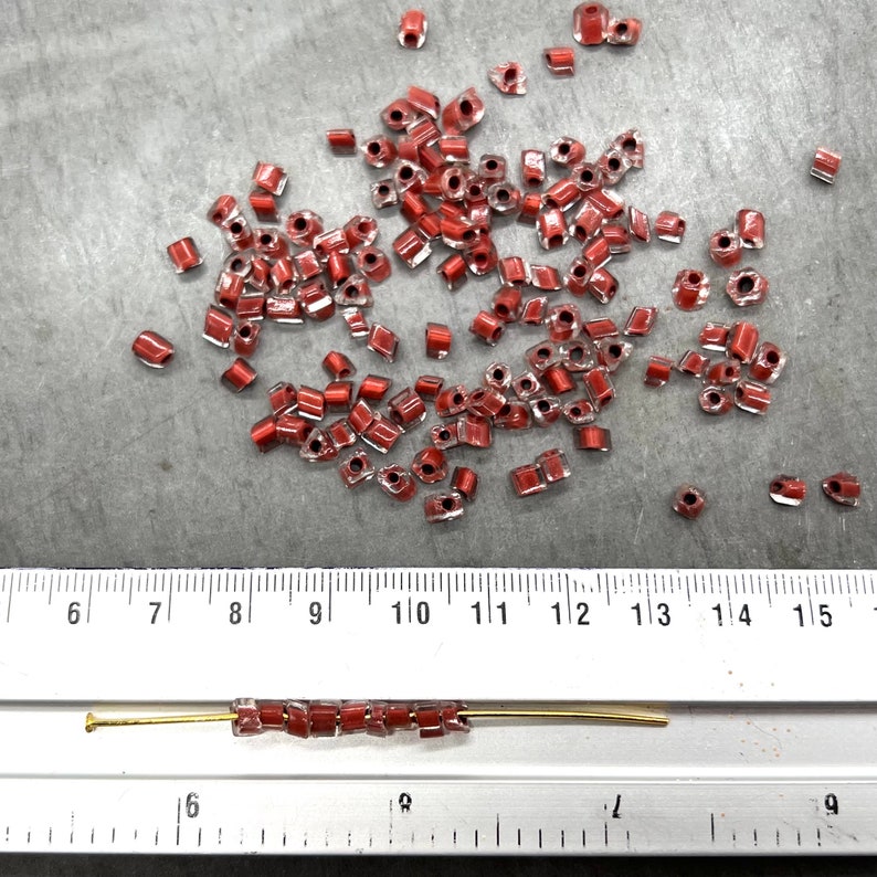 May include: A close-up of a ruler measuring 15 inches, with a line of small, red, square-shaped glass beads strung on a gold wire. The beads are scattered around the ruler.