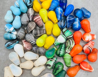 May include: A collection of colorful glass pear-shaped beads in various shades of blue, green, red, yellow, and white. The beads are arranged in a circular pattern on a gray surface.