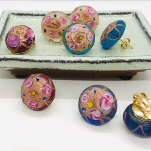 May include: A set of six colorful glass earrings with gold accents and pink and blue floral designs. The earrings are displayed on a light green ceramic tray.