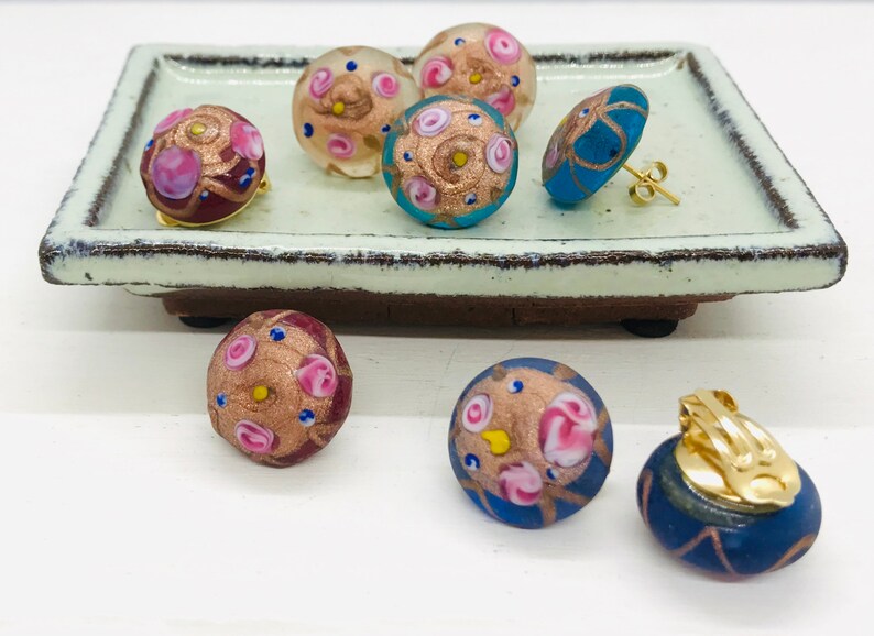 May include: A set of six colorful glass earrings with gold accents and pink and blue floral designs. The earrings are displayed on a light green ceramic tray.