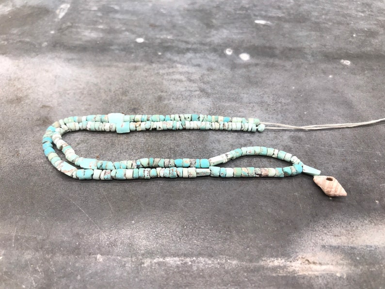 K&ouml;nnte beinhalten: Eine t&uuml;rkisfarbene Perlenkette mit einem Muschelanh&auml;nger. Die Perlen sind eine Mischung aus hell- und dunkelblau.