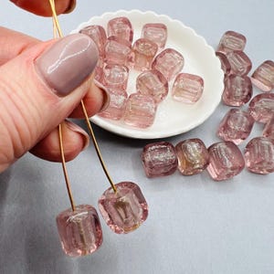 Può includere: Una collezione di perline di vetro rosa con una finitura scintillante. Le perline sono di forma quadrata e hanno un foro al centro per l'infilaggio. Le perline sono esposte su un piatto bianco e alcune sono tenute da una mano.