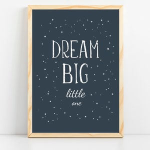 以下が含まれることがあります： ネイビーブルーの背景に「DREAM BIG little one」と書かれた白い文字がプリントされた額入りのプリント。小さな白い星が背景に散りばめられています。フレームは明るい色の木材で作られています。