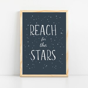 以下が含まれることがあります： 濃い青色の背景と白い星が描かれた額装されたプリント。プリントには、白い手書きのフォントで「REACH for the STARS」というテキストが書かれています。