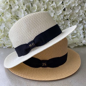 Puede incluir: Dos sombreros fedora de paja apilados verticalmente. El sombrero superior es de color crema con una banda negra que presenta las iniciales doradas H.G. El sombrero inferior es de color tostado con una banda negra y las iniciales doradas JS. Textura tejida.