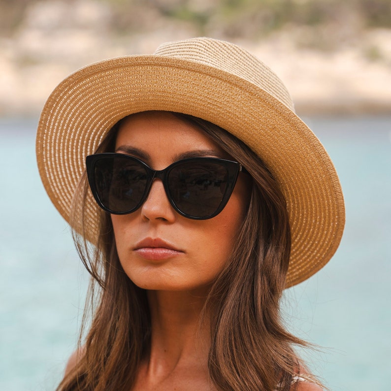 Puede incluir: Un sombrero fedora de paja color canela con ala ancha y textura tejida, combinado con gafas de sol negras estilo ojo de gato con lentes oscuras. El sombrero tiene una copa estructurada y un patr&oacute;n de tejido consistente en todo el material.