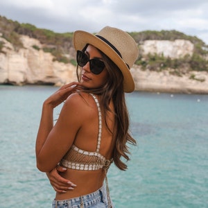 Puede incluir: Un sombrero fedora de paja beige con una cinta negra, combinado con un top bralette de ganchillo blanco y dorado met&aacute;lico con tirantes finos y cierre de lazo en la espalda, usado con pantalones cortos de mezclilla azul claro y gafas oscuras.