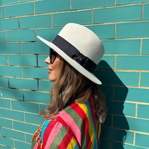 Puede incluir: Un sombrero fedora de paja color crema con una banda ancha de cinta de grosgrain negra alrededor de la copa. La cinta incluye las letras EW bordadas en una fuente sutil. El sombrero tiene un ala estructurada y una textura tejida.