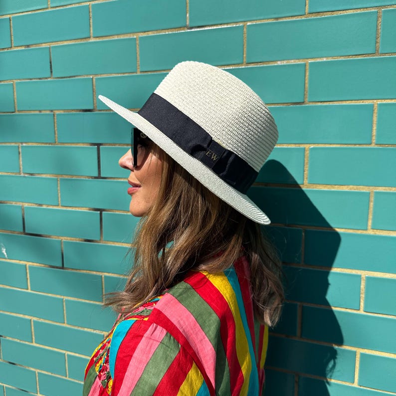 Puede incluir: Un sombrero fedora de paja color crema con una banda ancha de cinta de grosgrain negra alrededor de la copa. La cinta incluye las letras EW bordadas en una fuente sutil. El sombrero tiene un ala estructurada y una textura tejida.