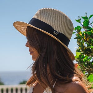 Puede incluir: Un sombrero fedora de paja beige con una banda de cinta negra ancha alrededor de la copa. La cinta negra est&aacute; decorada con las letras doradas SG en el lateral. El sombrero tiene un ala estructurada y una textura tejida en todo el material.