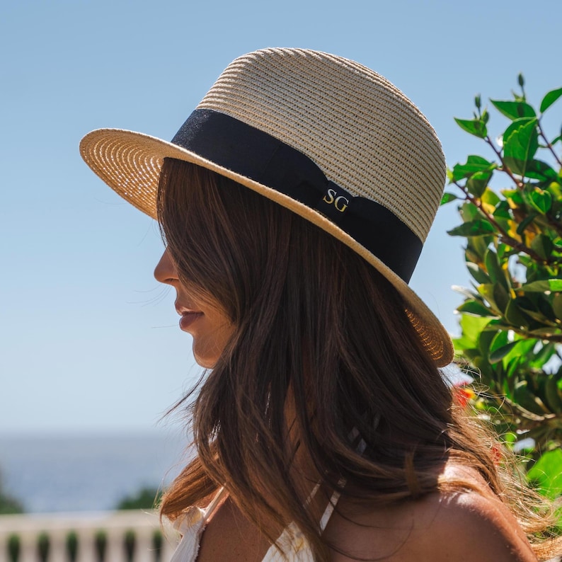 Puede incluir: Un sombrero fedora de paja beige con una banda de cinta negra ancha alrededor de la copa. La cinta negra est&aacute; decorada con las letras doradas SG en el lateral. El sombrero tiene un ala estructurada y una textura tejida en todo el material.